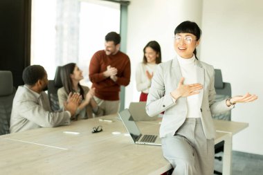 Güneşli bir ofiste canlı tartışmalar yapan çeşitli profesyonellerden oluşan bir grup. Bir kadın, dinamik bir çalışma ortamında takım çalışması ve işbirliği sergileyerek kendinden emin duruyor..
