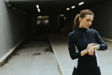 Şık bir şehir ortamında, odaklanmış bir kadın spor izleyicisini kontrol eder, canlandırıcı bir sabah egzersizi için hazırlanır..
