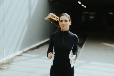 Kararlı bir kadın şık bir şehir koridorunda koşar, güneş ışığı içeri akar ve ayaklarının altındaki serin zeminde dinamik gölgeler bırakır..