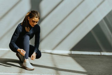 Bir kadın pürüzsüz bir yüzeye çömelir, ayakkabı bağcıklarını, gölgelerden akan güneş ışığıyla ayarlar, aktif bir güne hazırlanır..