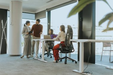 Meslektaşlar parlak, geniş bir ofis içinde işbirliği içinde toplandılar. Fikirlerin buluşması, bir üyenin masalarında odaklanmış takım arkadaşları tarafından çevrelenen sezgileri paylaşmasıyla gelişir..