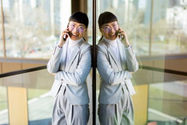 Modern ofis ortamında, şık takım elbiseli kendine güvenen Asyalı bir kadın telefonda konuşurken geniş bir gülümseme sergiliyor, yansıması şehrin cam zemininde görülebiliyor..