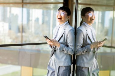 Şık gri takım elbiseli neşeli, profesyonel bir Asyalı kadın telefonunu tutarken gülümsüyor. Geniş pencereli modern bir ofiste duruyor. Dışarıdaki canlı şehri yansıtıyor..