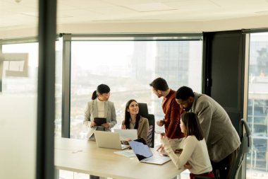 Bir grup profesyonel, geniş pencereleri ve doğal ışığı olan çağdaş bir ofiste proje planlarını aktif olarak tartışıyor. Müdahaleleri takım çalışması ve yenilik sürecini yansıtıyor.