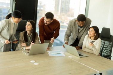 Çeşitli profesyonellerden oluşan bir grup, şık bir ofisteki masanın etrafında iş birliği yaparak, dizüstü bilgisayarlardan yararlanarak fikir ve içgörülerini paylaşıyor. Atmosfer canlı ve çekici..