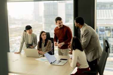 Bir grup profesyonel, geniş pencereleri ve doğal ışığı olan çağdaş bir ofiste proje planlarını aktif olarak tartışıyor. Müdahaleleri takım çalışması ve yenilik sürecini yansıtıyor.