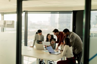 Bir grup profesyonel, geniş pencereleri ve doğal ışığı olan çağdaş bir ofiste proje planlarını aktif olarak tartışıyor. Müdahaleleri takım çalışması ve yenilik sürecini yansıtıyor.