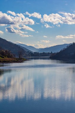 Parlak mavi bir gökyüzünün altında, gür yeşillik suyu sararken huzurlu bir göl kabarık bulutları yansıtır. Yumuşak tepeler ufku sarar, doğada sakin bir kaçış yaratır..