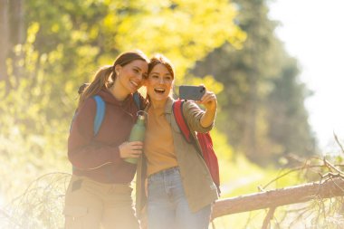 İki arkadaş güler ve bir selfie için poz verir. Sonbahar güneşinin ılık parıltısında canlı bir orman patikasını keşfederken..