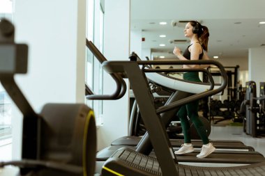 Parlak bir spor salonundaki koşu bandında odaklanmış bireysel egzersizler, kulaklıklar takılı, egzersiz ekipmanlarıyla çevrili..