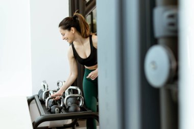 Gülümseyen kadın kettlebell eğitimini parlak, şık bir spor salonunda alıyor. Formuna ve sağlıklı yaşam tarzına bağlılığını gösteriyor..