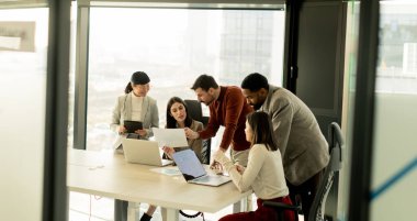Çeşitli profesyonellerden oluşan bir grup, bir konferans masası etrafında dinamik bir beyin fırtınası yapıyor, fikirlerini paylaşıyor ve parlak bir ofis alanında dizüstü bilgisayar ve notlarla etkileşime giriyor..