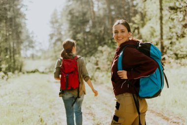 Dostlar, uzun ağaçlarla çevrili sıcak güneşin altında gülümseyerek ve açık hava maceralarının tadını çıkararak huzurlu bir orman yolunda ilerlerler..
