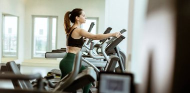 Çağdaş bir spor salonundaki parlak pencerelerle çevrili eliptik üzerinde odaklanmış bir egzersiz yapan bir kadın..