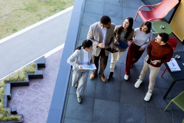 Beş profesyonelden oluşan çeşitli bir grup, lüks bir terasta dijital tabletleri incelerken canlı tartışmalara giriyorlar. Güneş ışığı kıyafetlerine neşe saçar..