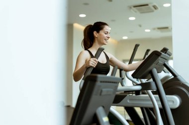 Bir kadın, iyi ışıklandırılmış bir spor salonunun enerjik atmosferine dalmış eliptik bir makinede egzersiz yapmaktan zevk alır..