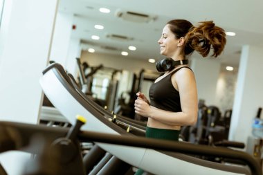 Neşeli bir kadın koşu bandında sürekli koşuyor. Parlak ve havadar bir spor salonunda, gelişmiş ekipmanlarla çevrili bir yerde antrenmanının keyfini çıkarıyor..