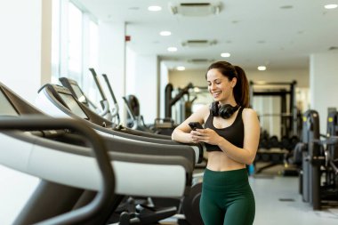 Günışığıyla dolu modern bir spor salonunda, genç bir kadın telefonunu kontrol ederken gülümsüyor, koşu bandında egzersiz yapmaya ara veriyor..