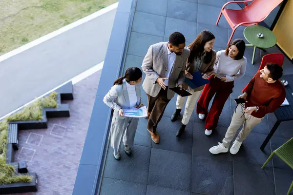 Beş profesyonelden oluşan çeşitli bir grup, lüks bir terasta dijital tabletleri incelerken canlı tartışmalara giriyorlar. Güneş ışığı kıyafetlerine neşe saçar..