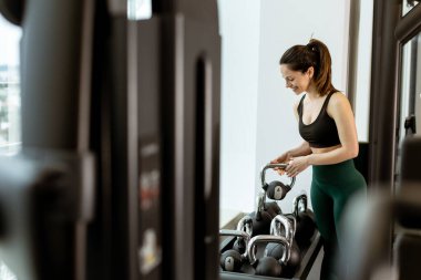 Odaklanmış bir birey, ışık dolu bir spor salonunda antrenman yaparken, çağdaş ekipmanların arasında kendini adama ve dayanıklılığı somutlaştırarak su ısıtıcılarını kaldırır..