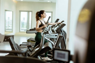 Kendini adamış bir kadın, büyük pencerelerle çevrili, parlak, çağdaş bir spor salonunda, eliptik bir makineyle fitness rejimine girer..