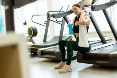 Yorgun ama tatmin olmuş bir kadın, elinde bir su şişesiyle koşu bandında rahatlar ve alnından ter silerken hak ettiği bir moladan zevk alır..