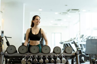 Kararlı bir kadın, parlak ve enerjik bir spor salonunda gücünü arttırmak için bir halterin yanında duruyor..