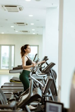 Doğal ışık dolu parlak bir spor salonu atmosferiyle çevrili eliptik bir makine kullanırken bireysel olarak fitness üzerine odaklanır..