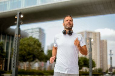 Kulaklıklı bir adam, güneşli bir sabahta çağdaş binalar ve yeşilliklerle çevrili verimli bir şehir parkında enerjik bir şekilde koşar..