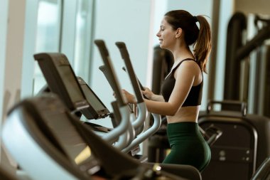 Genç bir kadın gündüz vakti şık bir spor salonunda eliptik bir makinede odaklanmış bir kardiyo çalışmasıyla meşgul oluyor..