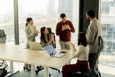 Bir grup profesyonel, geniş pencereleri ve doğal ışığı olan çağdaş bir ofiste proje planlarını aktif olarak tartışıyor. Müdahaleleri takım çalışması ve yenilik sürecini yansıtıyor.