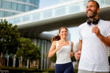 İki koşucu neşeli bir şehir parkında koşarken gülümsüyor. Etrafı parlak binalar ve güneşli bir sabahta verimli manzaralarla çevrili..