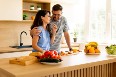 Sebze doğrayan ve doğal ışıkla dolu çağdaş bir mutfakta gülümseyen bir çift arasında neşeli bir etkileşim var..