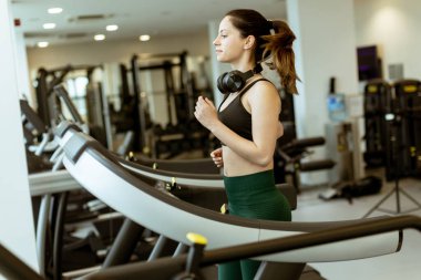 Fitness yolculuğuna odaklanmış genç bir kadın, modern bir spor salonundaki koşu bandında özenle koşuyor ve sabah rutinini kucaklıyor..