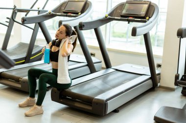 Yorgun ama tatmin olmuş bir kadın, elinde bir su şişesiyle koşu bandında rahatlar ve alnından ter silerken hak ettiği bir moladan zevk alır..