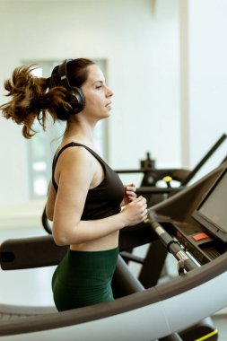 Bir kadın antrenmanından zevk alıyor, koşu bandına odaklanıyor, kulaklıklarını takıyor, doğal ışıkla dolu parlak bir spor salonunda enerji harcıyor..