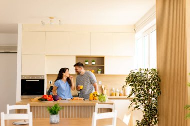 Sebze doğrayan ve doğal ışıkla dolu çağdaş bir mutfakta gülümseyen bir çift arasında neşeli bir etkileşim var..