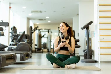 Kulaklık tutarken coşkuyla gülümseyen genç bir kadın, canlı bir spor salonunda bir anlık mutluluğun tadını çıkarıyor..