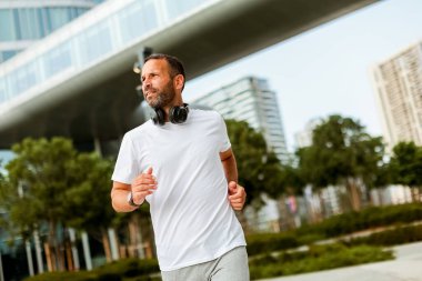 Kulaklıklı bir adam, güneşli bir sabahta çağdaş binalar ve yeşilliklerle çevrili verimli bir şehir parkında enerjik bir şekilde koşar..