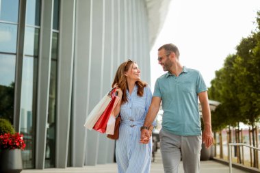 Neşeli bir çift, güneşli bir öğleden sonra, parlak bir mimari ve yemyeşil ortamda alışveriş çantalarına hayran bir şekilde el ele dolaşır..