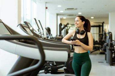Antrenman kıyafetleri içinde gülümseyen genç bir kadın koşu bandının yanında duruyor, ara veriyor ve modern bir spor salonunda telefonunu kaydırıyor..