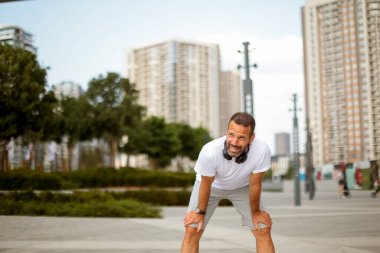 Yorgun ama mutlu bir adam açık gökyüzünün altında ağaçlarla ve çağdaş binalarla dolu canlı bir şehir parkında koştuktan sonra soluklanıyor..