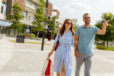 Bir çift el ele yürür, gelişigüzel giyinir, sıcak güneş ışığını emerler. Yeşillik ve çağdaş mimari ile dolu modern bir şehir alanını keşfederken alışveriş torbaları taşıyorlar..
