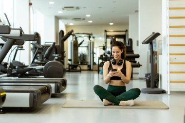 Gülümseyen kadın yoga minderinde kulaklıklarla oturuyor, canlı ve iyi donanımlı bir spor salonunda telefonuna odaklanmış..