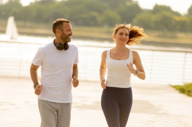 Bir çift, sıcacık sabah güneşinin tadını çıkararak, deniz manzaralı bir patika boyunca birlikte koşuyor. Gülüşleri aktif olmanın ve doğanın güzelliğinin tadını çıkarmanın neşesini yansıtıyor..