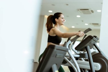 Bir kadın, eliptik bir makinede canlı bir egzersiz yapar. Sabahları aydınlık bir spor salonunda mutluluk saçar..