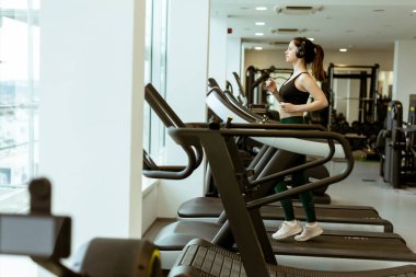 Kararlı bir kadın koşu bandında koşar. Parlak bir spor salonunda gelişmiş egzersiz ekipmanlarıyla birlikte sabah sporunun keyfini çıkarır..