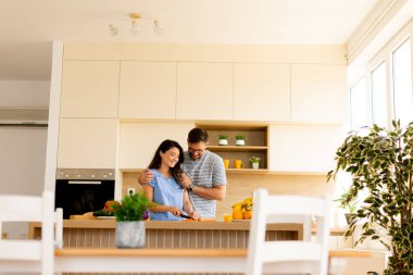 Bir çift, şık, güneşli mutfaklarında bitkiler ve canlı malzemelerle dolu bir yemek hazırlarken keyifli bir anı paylaşıyor. Kahkaha ve arkadaşlık havayı doldurur..