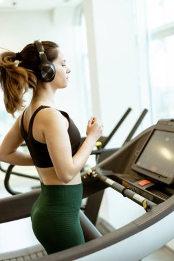 Bir kadın antrenmanından zevk alıyor, koşu bandına odaklanıyor, kulaklıklarını takıyor, doğal ışıkla dolu parlak bir spor salonunda enerji harcıyor..