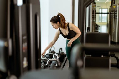 Çağdaş bir spor salonunda ağırlık seçen bir kadın, ilham verici bir çevrede güç ve sağIık kazanmaya odaklanır..